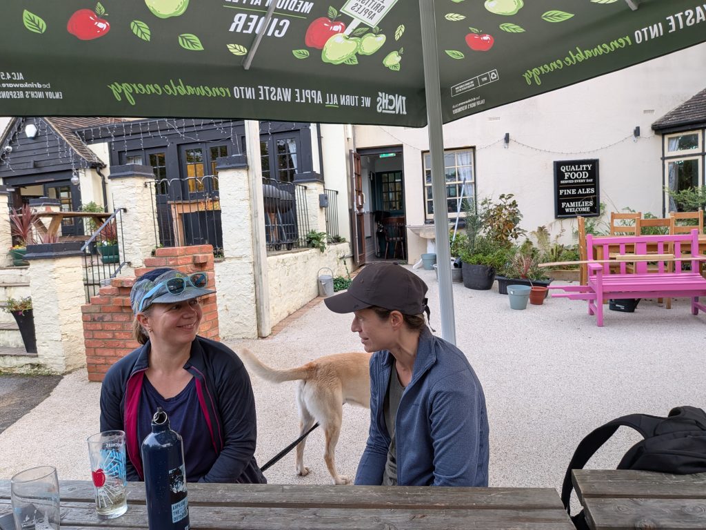 Syla and a friend chatting at an outdoor pub picnic table shaded by a green cider umbrella, with yellow Labrador Poppy standing in the background near a bright pink wooden bench.