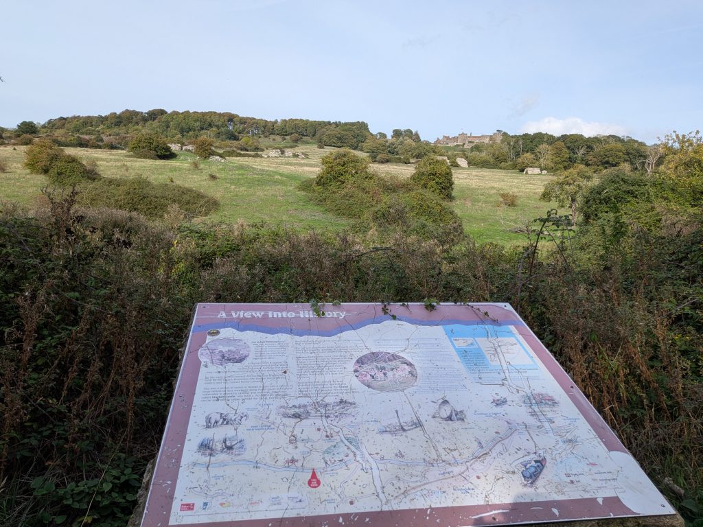 An illustrated heritage map board titled 'A View Into History' overlooking a vast green valley, with the stone ruins of Lympne Castle visible on the distant wooded ridgeline.
