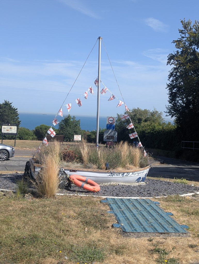 A decorative white boat named 'Spirit of St Margaret's' filled with coastal grasses and adorned with St George's Cross flags, sitting above the English Channel.