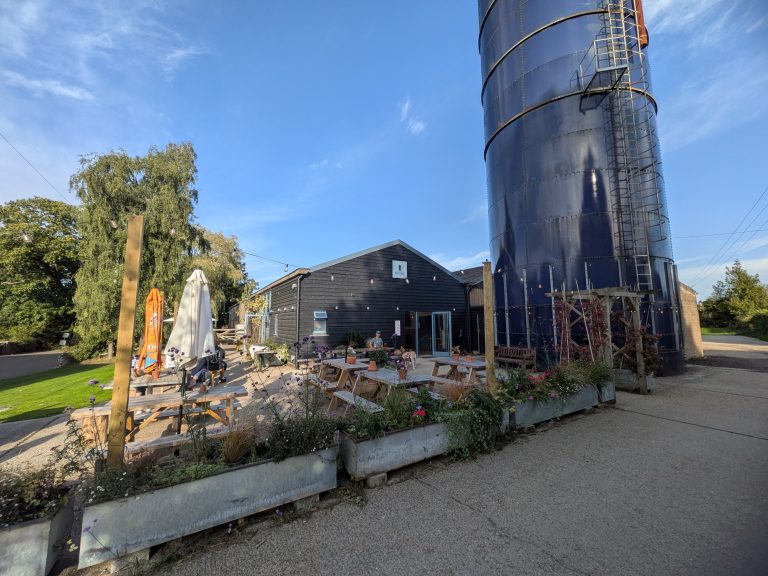 Outdoor beer garden patio at The Blue Silo with wooden picnic tables, flower planters, and the tall blue silo on a sunny day