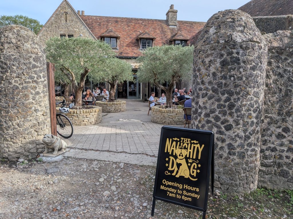 A black sandwich board sign for 'The Naughty Dog' cafe placed in front of a stone building courtyard featuring outdoor seating and olive trees in circular stone planters.