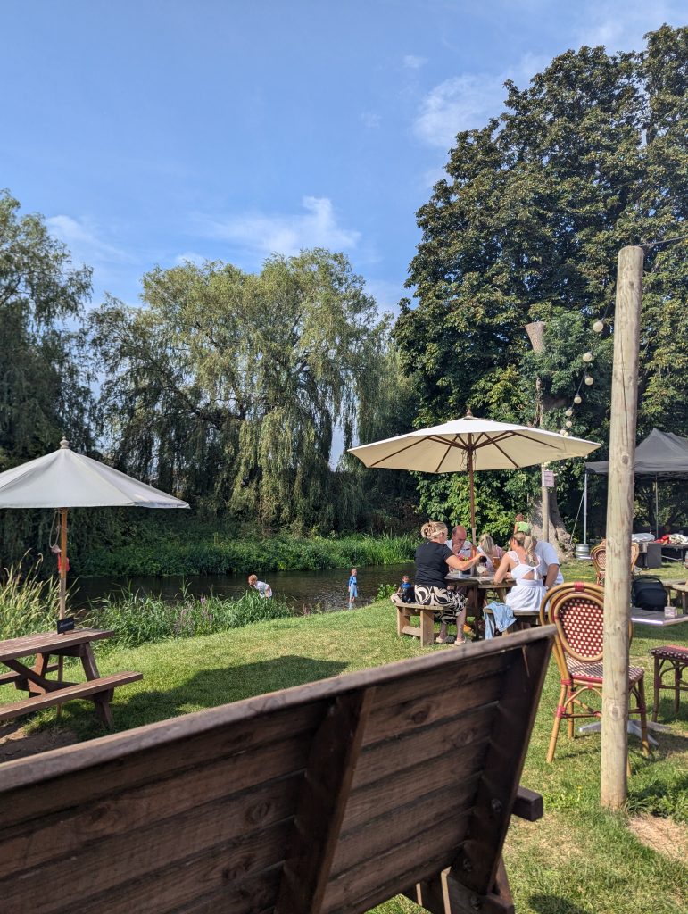 Patrons enjoying food and drinks at picnic tables with umbrellas in The Tickled Trout's riverside beer garden, while children play in the shallow river.