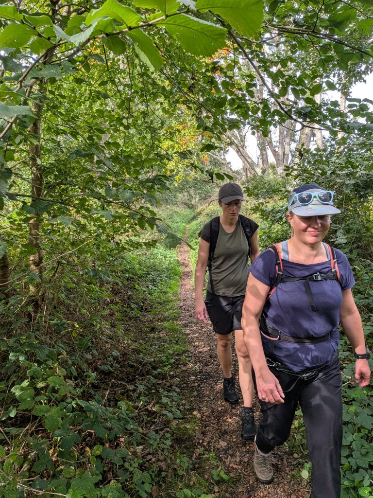 Syla and a fellow hiker walking (Aga) along a narrow, leaf-strewn woodland trail bordered by dense green foliage.