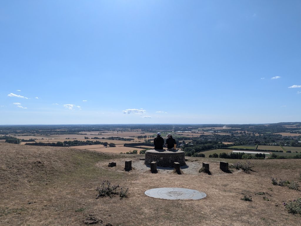 Two hikers resting on the Wye Crown stone marker looking out over the vast agricultural landscape of the Weald of Kent.